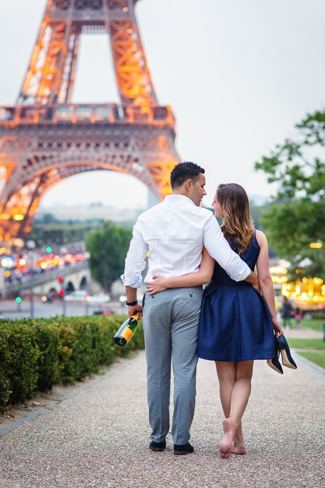 Summer engagement photos - Engaged couple walking towards the Eiffel Tower with shoes and champagne in hand Summer engagement photos in Paris - Engaged couple walking towards the Eiffel Tower with shoes and champagne in hand